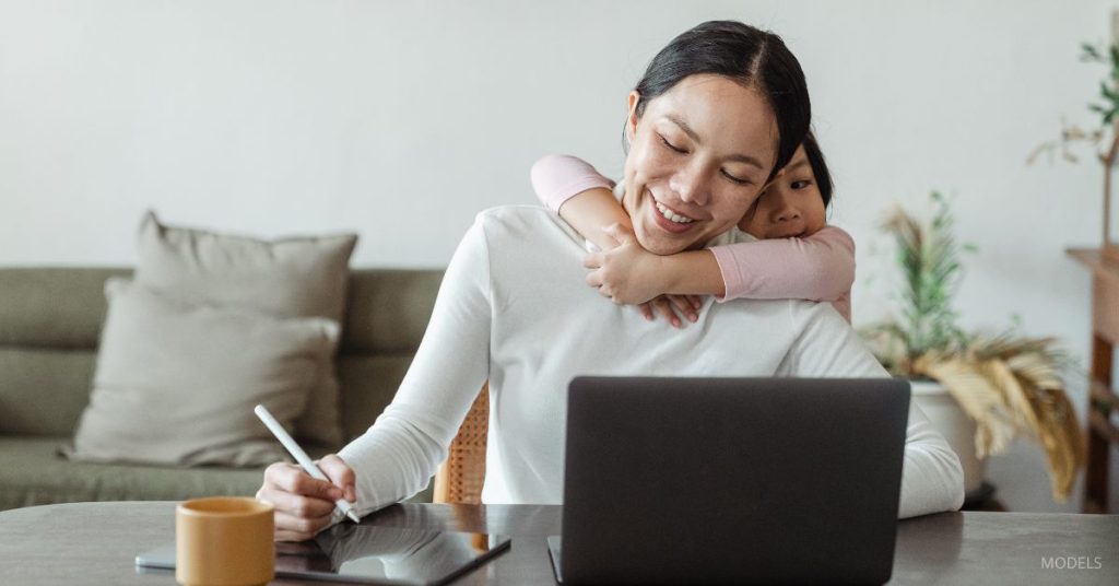 Woman sitting at a computer with her daughter wrapped around her shoulders (models) working and smiling indoors.