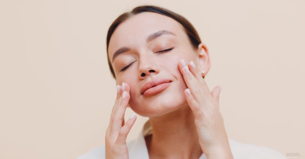A women's face is shown with her hair pulled back, eyes closed, and her hands gently touching the sides of her faces. (model)